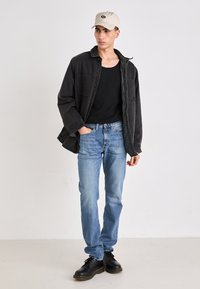 Zwarte denim oversized shirt, aansluitend zwart tanktop, lichtblauwe jeans en zwarte veterschoenen. Draagt een beige pet met een logo.