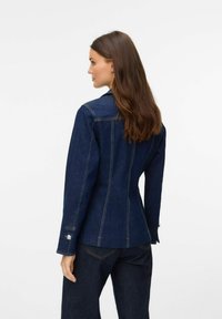 Tumedast sinine denim jakk, millel on kohandatud lõige, kontrastne õmblus ning nööbid mansettidel. Omab klassikalist kraed ja seljatükki kaunistavat detaile.