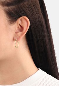 Närbild på en persons vänstra öra med långt brunt hår, iförande en liten snodd guldring och en vit stickad topp.