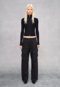 Top de manga larga negro acanalado con cuello alto y cremallera, combinado con pantalones cargo negros holgados que cuentan con grandes bolsillos laterales y zapatos planos.