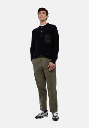 Pull en tricot noir avec une poche en patch, associé à un pantalon en velours côtelé olive. Les baskets présentent des accents gris, blanc et marine. Le modèle se tient avec confiance.