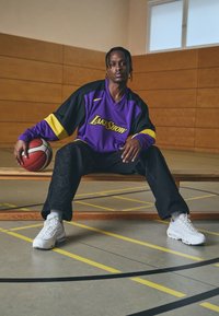 Violeta un melna pārsmagā džersija ar uzrakstu "LakeShow", kombinēta ar melnām biksēm ar faktūras rakstu un baltām kedām. Rokā tur basketbola bumbu.