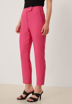 Frau trägt leuchtend pinke, maßgeschneiderte Knöchelhosen mit vorderem Verschluss, kombiniert mit schwarzen, offenen Sandalen mit Absatz und einem schwarzen ärmellosen Oberteil.