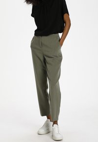 Pantalon vert olive clair à coupe ajustée, avec poches latérales et texture lisse, associé à un haut noir ample et des baskets blanches.