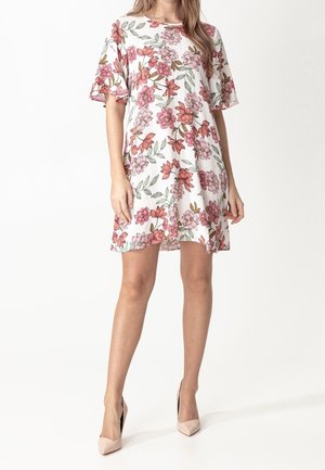 Femme portant une robe blanche à motifs floraux jusqu'aux genoux avec des manches volantées et des talons hauts couleur nude, debout devant un fond uni.