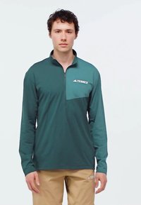 Dlhý rukáv, športová košeľa v odtieni teal s obojkom na zips, kontrastným predným panelom a vyšitým logom na ľavej strane. Hladká textúra.