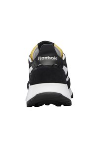 Svart og hvit idrettssko med semsket materiale, gule detaljer, tykk hvit såle og sølv Reebok-logo bak.