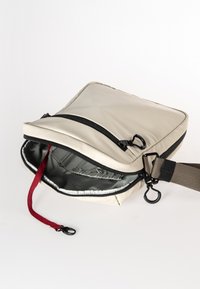 Beige schoudertas met een ritssluiting, een grijze binnenkant en een rode schouderband. Voorzien van zwarte hardware en een slank, gewatteerd ontwerp.