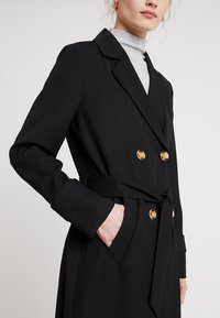 Kvinna i en svart dubbelknäppt jacka med guldknappar över en ljusgrå polotröja, handen i jackfickan, på en plain ljus bakgrund.