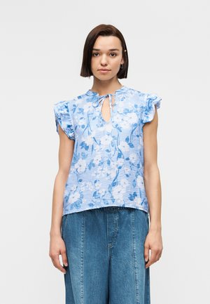 Žena oblečená v svetlomodrej kvetinovej blúzke bez rukávov s volánikovými ramenami a výstrihom na gombík, v kombinácii s voľnými modrými rifľovými nohavicami.