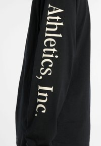 Musta pitkähihaine t-paita, joka on valmistettu puuvillasta. Hihoissa on valkoista tekstiä, jossa lukee "Athletics, Inc.", ja siinä on rento leikkaus.