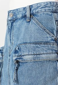 Svetlo modri jeans blizu, ki prikazuje sprednji žep, zanko za pas, kovinski gumb in podrobnost zadrževalnega žepa.