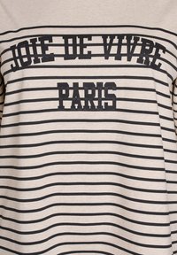 Béžová a černá pruhovaná látka s tučným černým textem "JOIE DE VIVRE" nad "PARIS" v blokovém písmu.