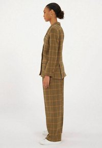 Costume à carreaux dans des tons marron et jaune, comprenant une veste ajustée et un pantalon large, fabriqué dans un tissu léger. Baskets blanches.
