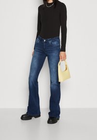 Dunkelblaue ausgestellte Jeans mit klassischem Fünf-Taschen-Design, kombiniert mit einem schwarzen Langarmtop und einer kleinen gelben Handtasche. Schwarze Plateauschuhe.
