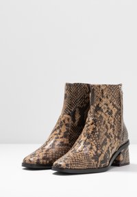 Un par de botas de tobillo con un patrón de serpiente en tonos marrón y negro, punteras puntiagudas, tacones cuadrados y cremalleras laterales sobre una superficie blanca.