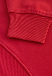 Červená mikina s žebrovanými manžetami a hladkou texturou. Má šitý šev v odpovídající červené barvě, který zdůrazňuje jednoduchý, jednotný design.