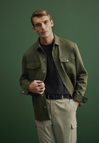 Chaqueta texturizada verde oliva con bolsillos frontales, camiseta negra debajo y pantalones beige. El modelo sostiene la chaqueta y lleva anillos de plata y un cinturón.