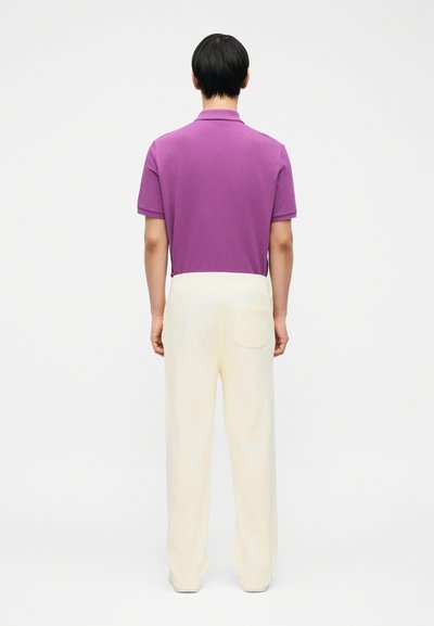 Άτομο που στέκεται με την πλάτη προς τα εμάς, φοράει μοβ πόλο μπλουζάκι με κοντά μανίκια και εκρού χαλαρή παντελόνα με τσέπη στο πίσω μέρος.