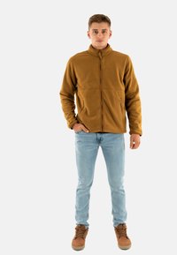 Chaqueta de felpa marrón con un cuello alto, cremallera frontal y dos bolsillos laterales. Combinada con jeans de color azul claro y botas marrones con cordones.