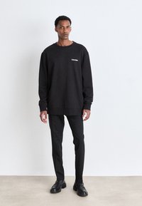 Sweatshirt noir avec col rond et détail logo, associé à un pantalon noir slim et des mocassins noirs. Tissu lisse ; design minimaliste.