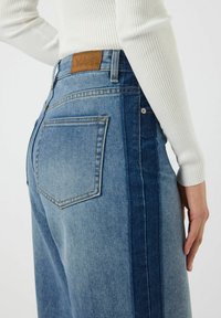 Jeans de mezclilla azul claro con un diseño de pierna ancha, una parche de cuero marrón con "YAS" y un panel lateral en un tono azul más oscuro.