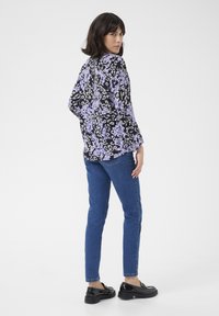 Blouse à motifs léopard noir et violet à manches longues, associée à un jean skinny bleu et des chaussures noires vernis. Fond minimaliste.