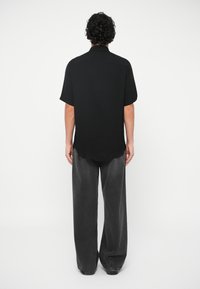 Μαύρο κοντομάν�ικο πουκάμισο με ελαφρώς κυρτή ραφή στο κάτω μέρος, συνδυασμένο με φαρδιά, φθαρμένα γκρι παντελόνια. Και τα δύο ρούχα είναι απλά και ουδέτερα.