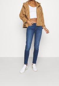 Beige waterproof jacket, white cropped tank, blue skinny jeans, and white sneakers with black accents. Casual outdoor outfit.