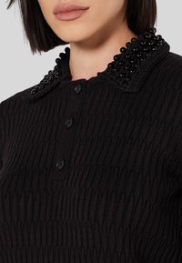 Top nero a coste con collo decorato da perline nere, quattro bottoni frontali e tessuto testurizzato.