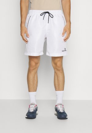 Mężczyzna ubrany w białe szorty ze sznurkiem, czarną lamówką i logo, białe skarpetki oraz granatowe buty FILA, stojący na białym tle.