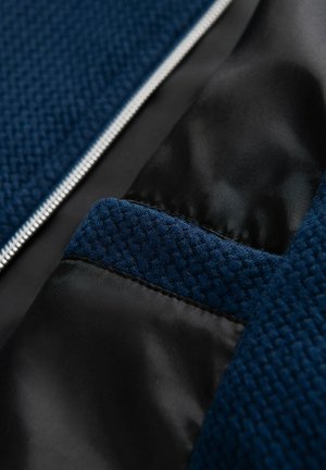 Mörkblå texturerad tyg med ett vävt mönster, med en slät svart satinfoder och en silverfärgad dragkedja längs en kant.