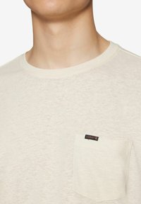 T-shirt a maniche corte color crema realizzato in un tessuto texturizzato, con una tasca a toppa con etichetta sul lato sinistro del petto. Collo rotondo.