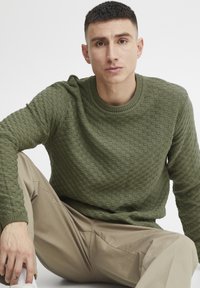 Olijfgroen gebreid sweater met een textuur patroon, ronde hals, ribbels aan de manchetten, gedragen met beige broeken. Model zit, hand rustend op de knie.