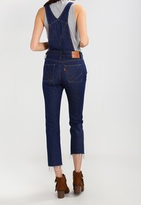 Woman wearing dark denim overalls, gray sleeveless top, and brown ankle boots, standing with back facing the viewer on a plain background.