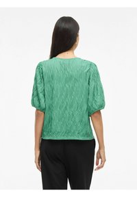 Blusa verde, con texture e un motivo ondulato, maniche a puff corte e scollo rotondo, abbinata a pantaloni neri.
