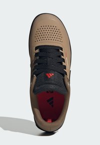 Ruskea urheilukenkä mustilla nauhoilla, rei'itetty kärki ja punainen sisus. Sileä materiaali, jossa on hienovarainen brändäys ja pyöreä muoto.