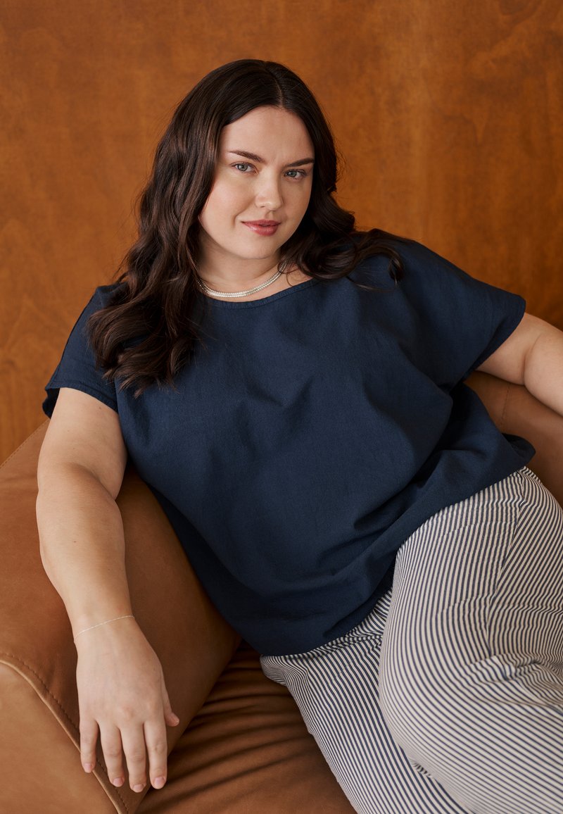 Femme aux longs cheveux bruns portant une chemise bleu marine et un pantalon rayé, allongée sur un canapé en cuir marron devant un fond en bois.