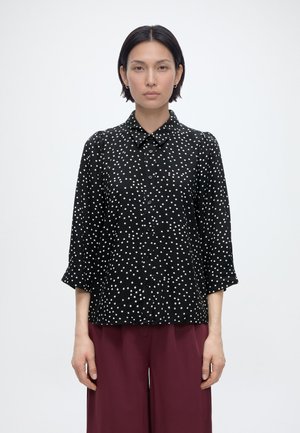 Kobieta o krótkich czarnych włosach ubrana w czarną, zapinaną na guziki bluzkę w białe kropki oraz luźne bordowe spodnie, stojąca na tle jednolitego jasnego tła.