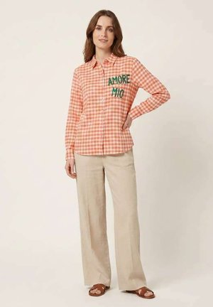 Femme portant une chemise à carreaux pêche avec boutons et texte "AMORE MIO", pantalon large beige et sandales marron, debout avec une main sur la hanche.