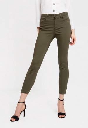 Mujer modelando pantalones ajustados de color verde oliva combinados con sandalias negras de tacón alto y una blusa blanca de manga larga con botones, corta a la cintura.