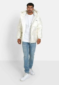 Chaqueta acolchada color crema con un alto cuello, grandes bolsillos frontales y costuras visibles. Lleva puesta sobre una camisa blanca y jeans rasgados de color azul claro, con zapatillas blancas.