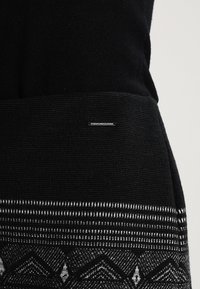 Schwarzer Pullover aus strukturiertem Strickmaterial. Verfügt über einen gemusterten Saum in schwarz-weißen geometrischen Designs. Kleine Markenetikette sichtbar.