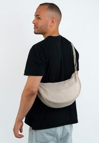Uomo che indossa una maglietta nera e jeans chiari porta a tracolla una borsa a tracolla beige Larkson sulla spalla, su uno sfondo bianco semplice.