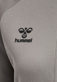 Grått tyg med ribbad textur, med en svart Hummel-logotyp och ett broderat bi-design framtill.