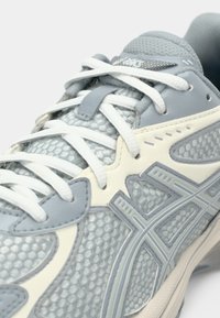 Gros plan d'une chaussure de sport grise et blanche avec des lacets blancs, un tissu en mesh et des superpositions synthétiques sur la partie supérieure.