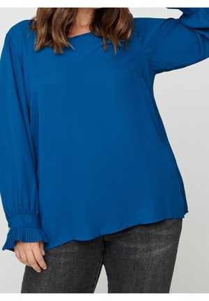 Blaue Langarmbluse mit lockerem Schnitt, rundem Ausschnitt und plissierten Manschetten, aus glattem Stoff; kombiniert mit dunkelgrauen Jeans.