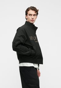 Jachetă bomber neagră cu guler înalt, având literă brodată și accente texturate pe mâneci. Țesătură netedă cu o croială lejeră.