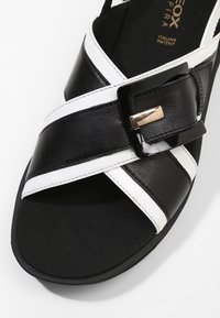 Mustavalkoinen nahkasandaali, jossa on ristikkäiset sangat ja musta solkiyksityiskohta. Pohja on musta ja siinä on teksturoitu pinta.