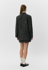 Melna un balta tvīda žakete ar garu siluetu, ar iegrieztu apkakli un strukturētu apdari, valkāta virs pieguļošām svārkiem un zeķēm.